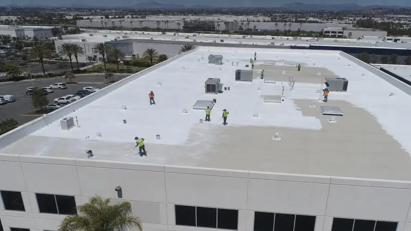 An aerial view of a large white-coated flat commercial roof with a crew in high-visibility vests rolling coating around the rooftop units, an industrial park and palm trees beyond