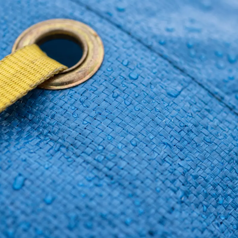 Close-up of a blue woven tarp beaded with rainwater, with a brass grommet and a yellow strap threaded through it